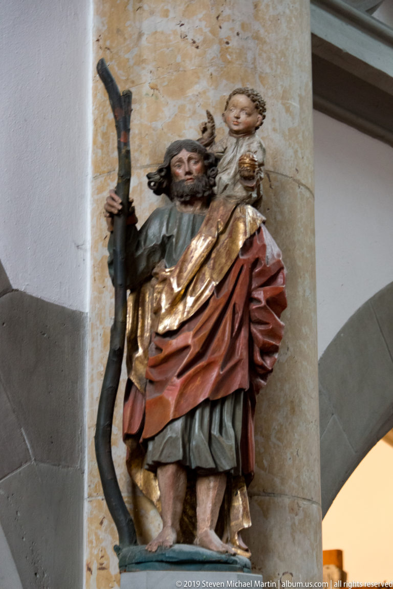 Polychrome Saint Christopher holding a staff and carrying the Christ Child on his shoulder beside a stone column.