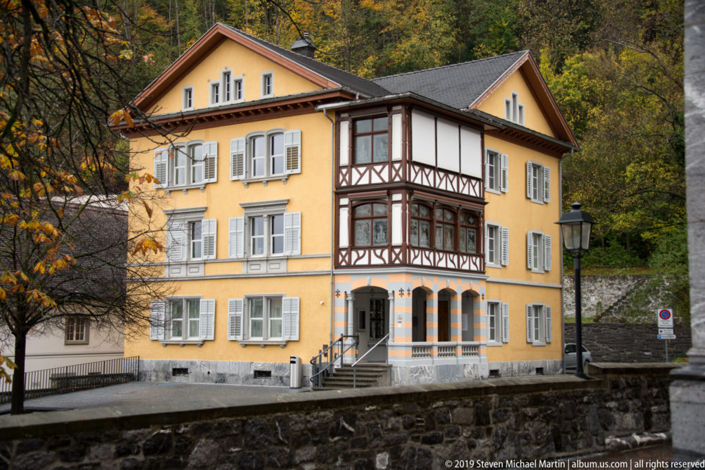 Frontal view of yellow house with gray shutters, projecting half-timbered bay, striped supports, and entrance steps.