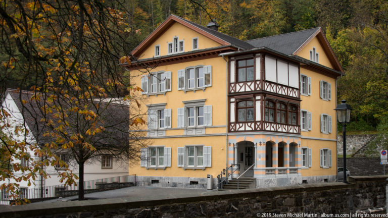 Yellow historic house with gray shutters and a tall dark half-timbered projecting bay, framed by autumn foliage.