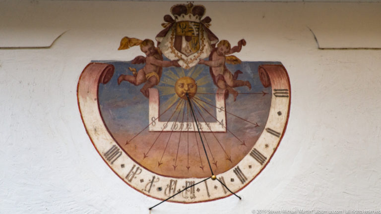 Painted wall sundial with sun face, hour lines, crowned coat of arms, two putti, and scroll-shaped border.