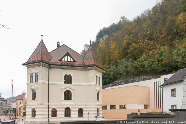 Historic cream building with patterned steep roofs and small turrets beside a pale modern civic building and wooded hillside.