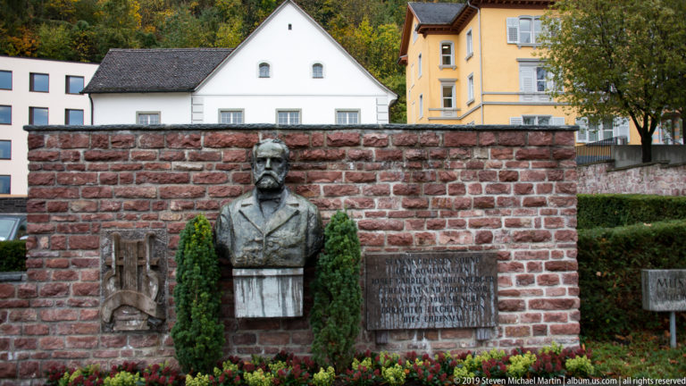 Bronze bust of a bearded man centered in a brick memorial wall with relief panels, plaque, shrubs, and flowers.