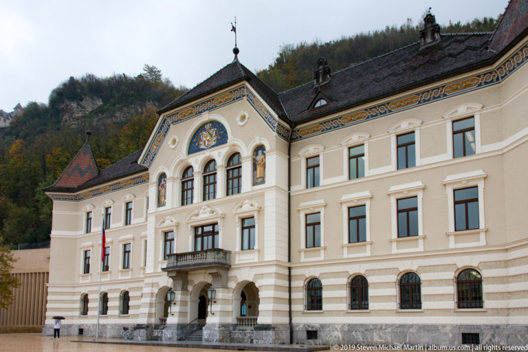Ornate Government Building in Vaduz with arched entry, central balcony, painted gable decoration, and patterned roof.