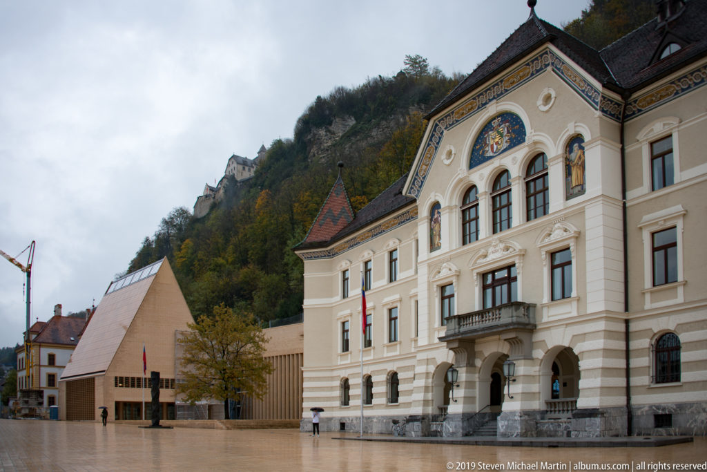 Historic Government Building beside the modern Landtag complex on a broad plaza, with wooded hillside and castle above.