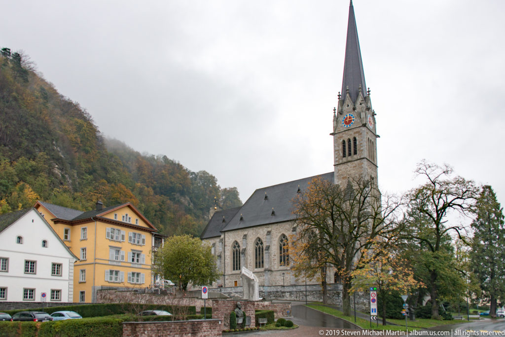 Cathedral of St. Florin in Vaduz with tall pointed spire, stone walls, Gothic windows, and autumn trees.