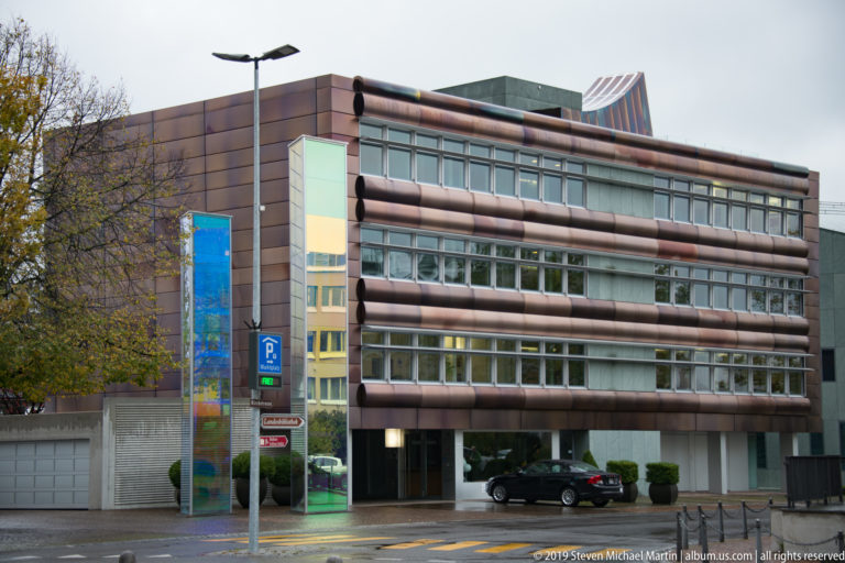 Modern multi-story office building with horizontal brown façade bands, broad windows, glazed corner bay, and parked cars.