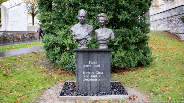 Full frontal view of the Franz Josef II and Princess Gina bust monument against a dense evergreen shrub.