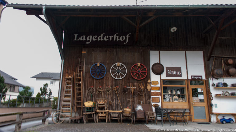 Timber Lagederhof façade with wagon wheels, farm tools, chairs, ladders, and metal vessels arranged around the entrance.