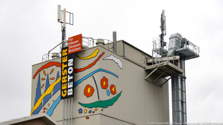Gerster Beton industrial building with colorful abstract mural, company sign, rooftop antennas, and metal service platforms.