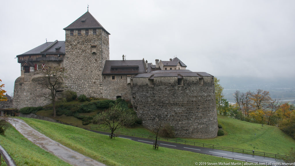 Side view of Vaduz Castle with square tower, stone walls, clustered roofs, wet grass, and misty valley beyond.
