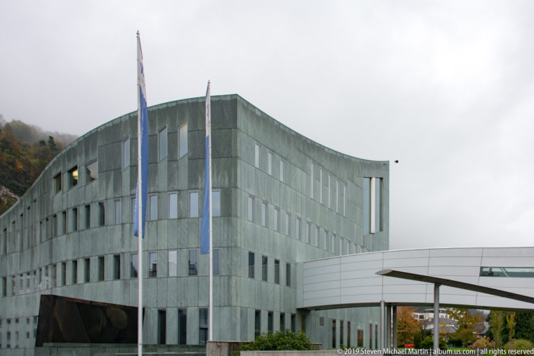 Curved green-gray stone building with rows of windows, three flagpoles, and a white connecting structure at right.