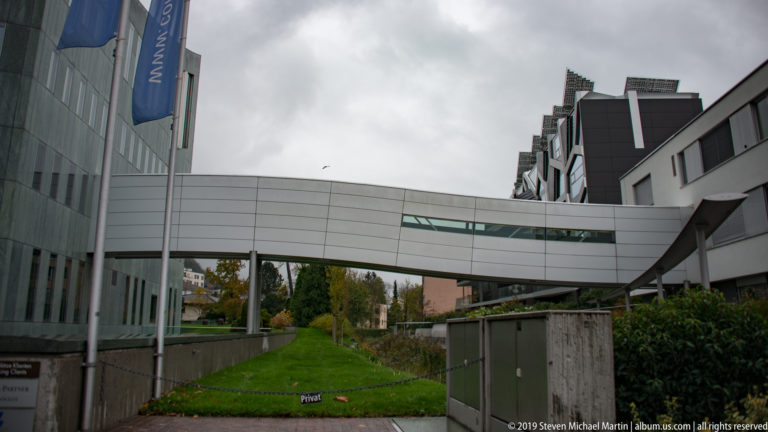 Enclosed white skybridge links modern buildings above a narrow landscaped passage under an overcast sky.