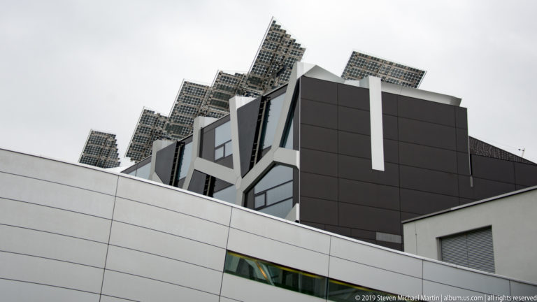 Low-angle view of a modern dark-and-white building topped by multiple tilted photovoltaic panel arrays.