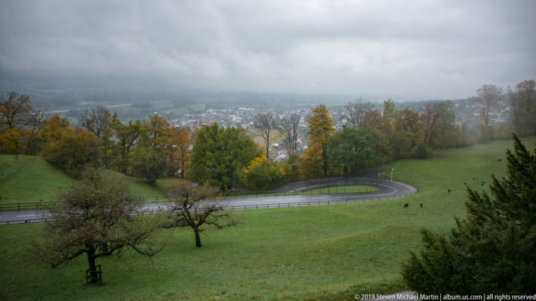Mist-covered valley view with green meadows, orchard trees, and a wet road curving down toward Vaduz.