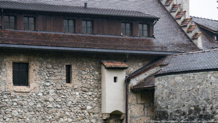 Close view of rough stone castle walls, narrow windows, tiled roofs, and dark timber upper cladding.