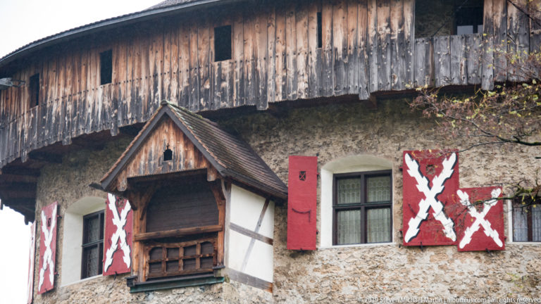 Weathered timber gallery above stone wall with small windows, patterned red shutters, and a gabled wooden projection.