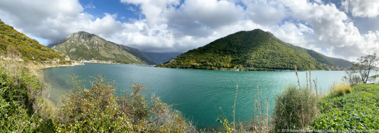 Turquoise Bay of Kotor curves between steep green mountains beneath bright clouds, with a road tracing the left shoreline.