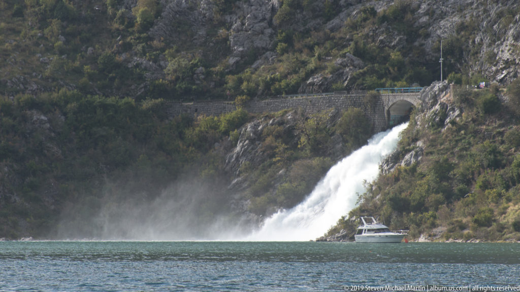 Sopot waterfall drops from a rocky slope toward Bay of Kotor water, with a small boat below and road bridge above.