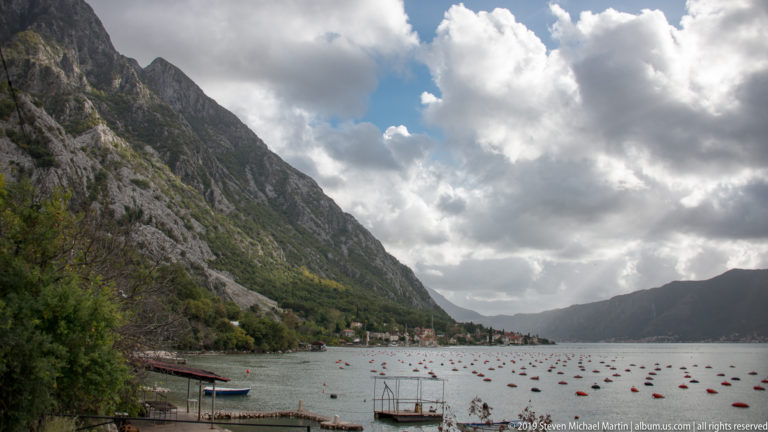 Rows of red and dark aquaculture floats cover calm Bay of Kotor water below a steep mountain and shoreline village.