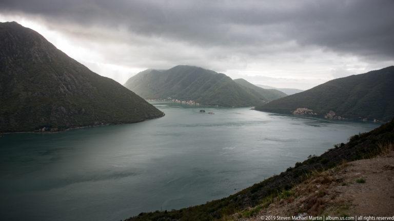 Bay of Kotor seen from above, with steep dark mountains, shoreline villages, and sheltered water beneath storm clouds.