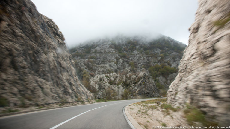 Curving paved road passes through narrow pale limestone cuttings toward a mist-covered rocky mountain slope.