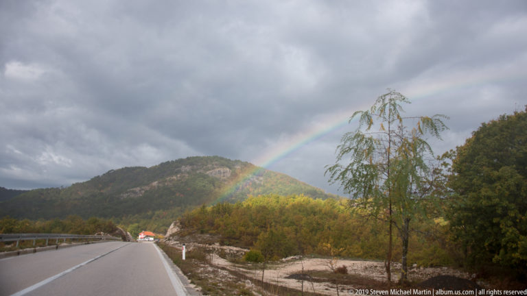 Rainbow descends toward an autumn-covered mountain beyond a two-lane highway under dark storm clouds.