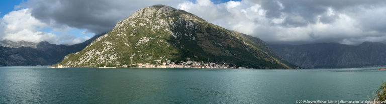 Perast forms a narrow waterfront strip beneath a steep green mountain across the blue-gray Bay of Kotor.