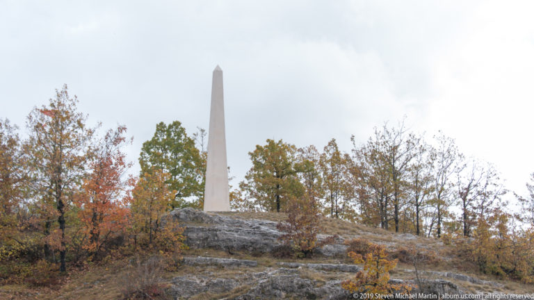 Slender pointed stone obelisk standing on exposed rock among autumn trees beneath a pale sky.