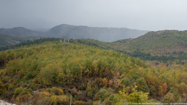 Misty mountain ridges behind broad autumn woodland in green, yellow, and rust tones under rain clouds.