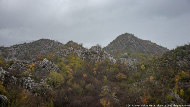 Jagged gray limestone ridge above subdued autumn woodland under a dark overcast sky in Montenegro.