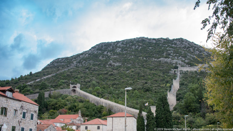 Long stone defensive wall climbs a steep green hillside above red-roofed houses in Montenegro.