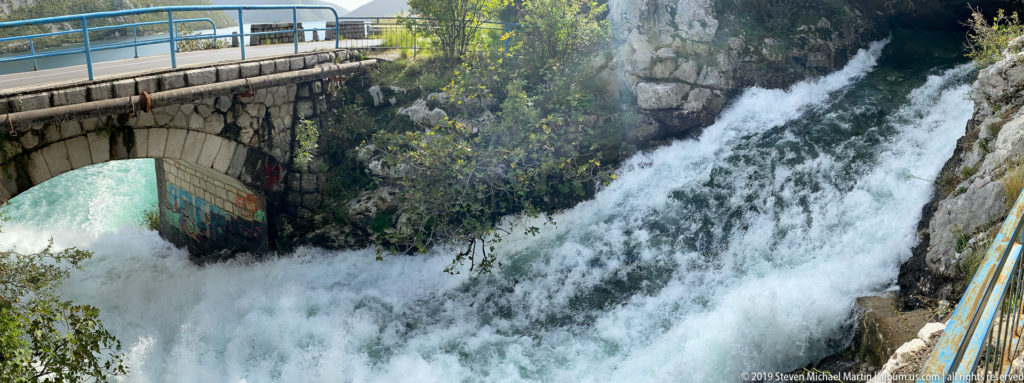 Foaming Sopot waterfall rushes beneath a stone-arched road bridge beside dark rock near Risan, Montenegro.