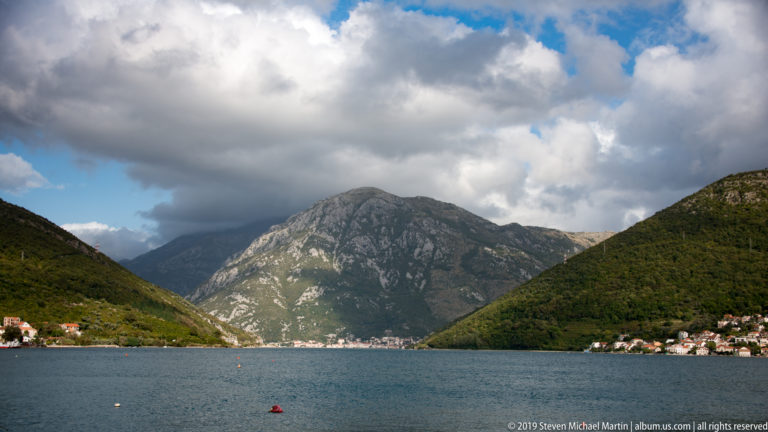 Wide Bay of Kotor view framed by steep green mountains, shoreline villages, calm water, and distant ridges.