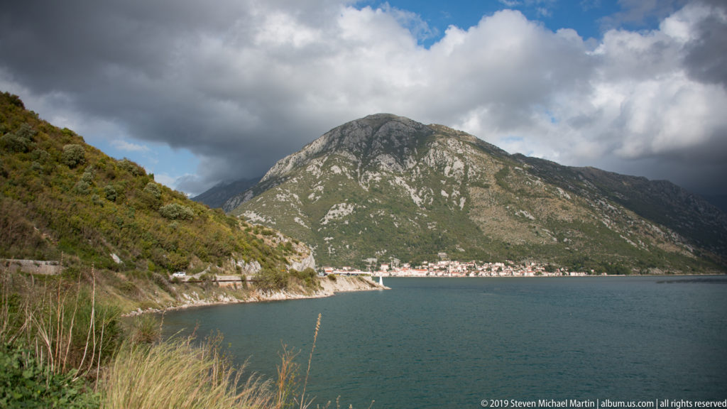 Rounded green limestone mountain rises behind a narrow Bay of Kotor waterfront settlement under large clouds.