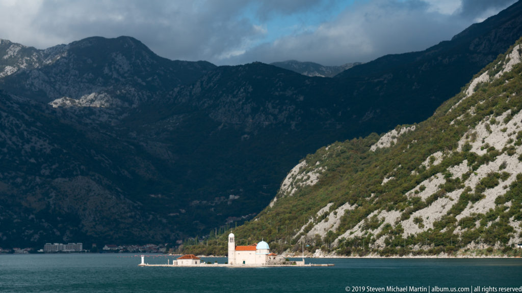 Our Lady of the Rocks island church with blue dome and bell tower beneath towering dark Bay of Kotor mountains.