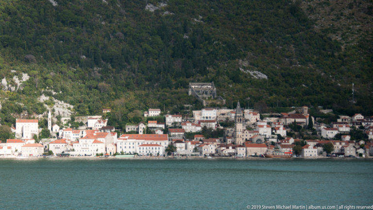 Perast waterfront with pale stone buildings, red roofs, and church towers beneath a steep forested mountain beside the Bay of Kotor.