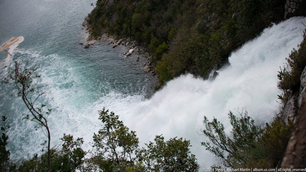 Foaming Sopot waterfall descends through a rocky, shrub-lined channel into turquoise water near Risan, Montenegro.