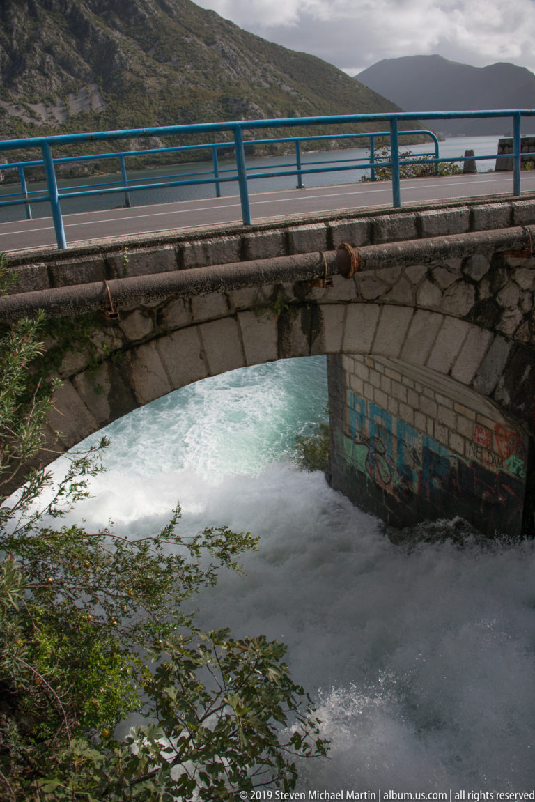 Foaming water surges beneath a stone-arched road bridge with blue railings near Risan on the Bay of Kotor.