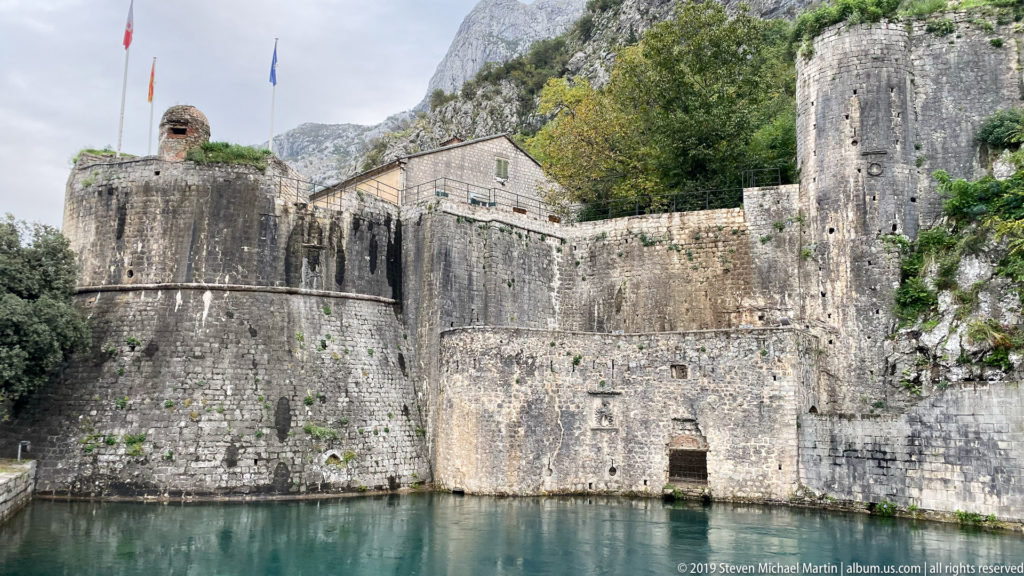 Rounded bastions and thick curtain walls of Kotor above water, with mountain vegetation and buildings behind.
