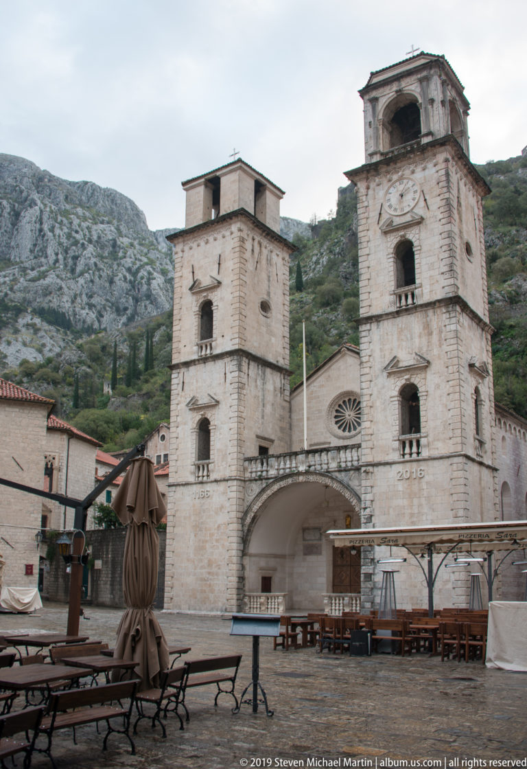 Lower façade and twin towers of St. Tryphon Cathedral behind café tables and folded umbrellas in Kotor.
