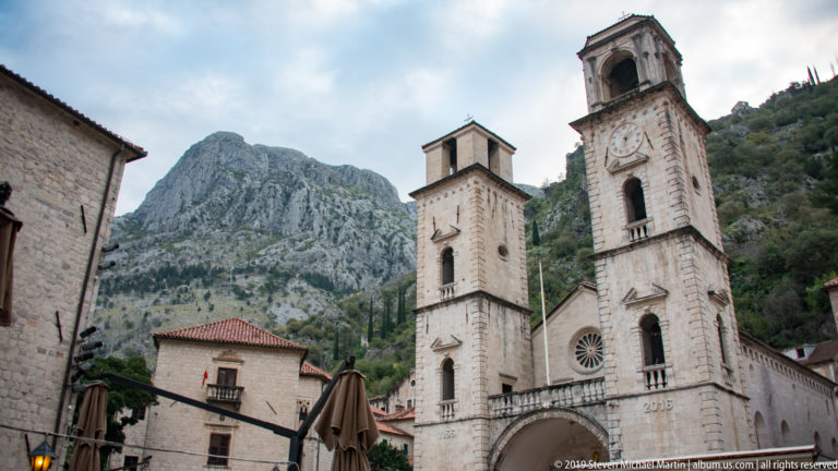 Cathedral of St. Tryphon across a paved square, with café umbrellas, neighboring buildings, and mountains behind.