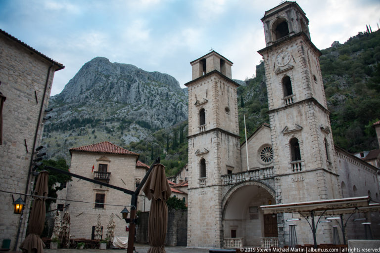 Cathedral of St. Tryphon with twin stone bell towers, rose window, arched entrance, and mountains behind.