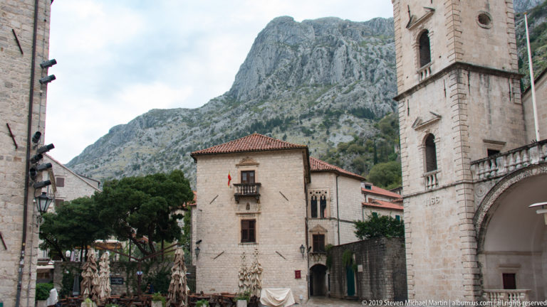 Partial twin-towered façade of St. Tryphon Cathedral beside historic stone buildings with steep mountains behind.