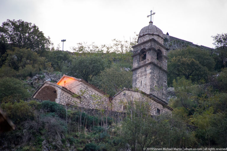 Small stone church with a square bell tower and cross on a steep wooded hillside above Kotor.