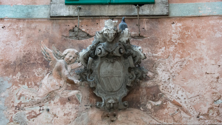 Stone heraldic cartouche with scrollwork and winged putti mounted on a weathered pink plaster façade.