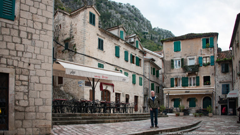 Historic stone buildings with green shutters and café seating beneath a steep rocky mountain in Kotor.