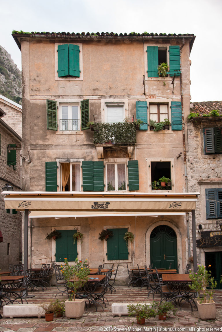 Weathered multi-story façade with green shutters, flower boxes, café awning, and outdoor tables in Kotor.
