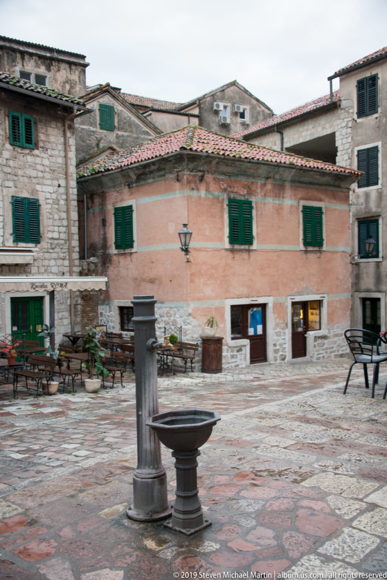 Small stone-paved square in Kotor with a central fountain, café seating, and historic buildings with green shutters.