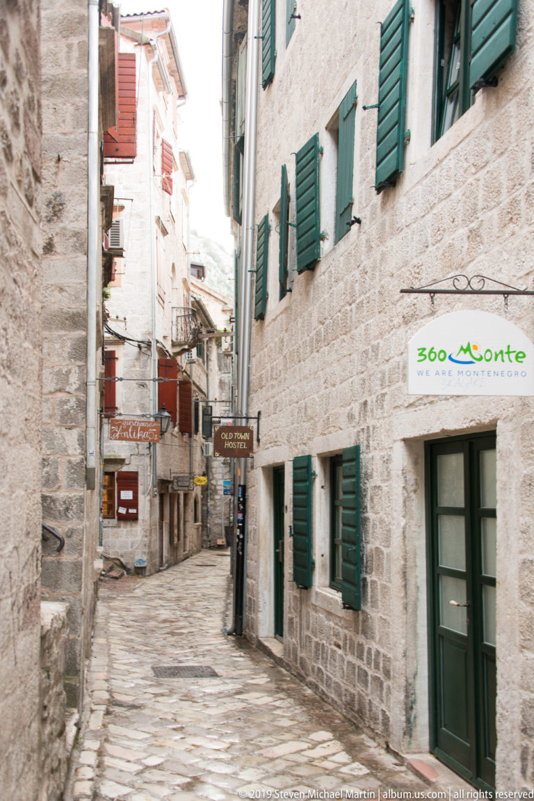 Narrow stone-paved lane in Kotor with pale masonry buildings, green shutters, shop signs, and overhead cables.