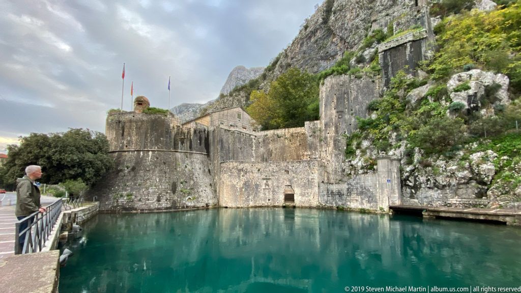 Massive Kotor city walls and rounded bastion above blue-green water, with a visitor at a railing and mountains behind.